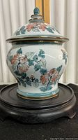 Porcelain urn with lid showing peach flowers and green leaves on white background, gold trim accents