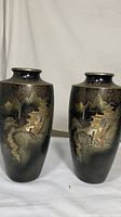 Pair of tall bronze vases with detailed etched and inlaid gold-toned landscape scenes of mountains, trees, and buildings on dark patina background. Both vases shown side by side.