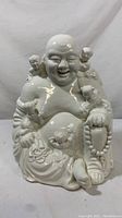 Front view of the white porcelain Hotei Buddha statue with children surrounding it showing detailed facial expressions and beads in hand.