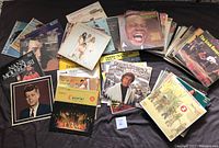 Wide view of the full lot of 67 vinyl records spread on a dark cloth background, showing variety of album covers including music and speech albums, classic and vocal artists.