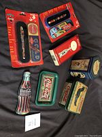 Photo showing two Coca-Cola branded pens in original packaging, one set with two pens, a boxed pizza cutter, a Coca-Cola shaped tip tray, and three metal Coca-Cola tins with vintage-style advertising.