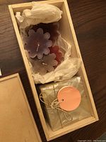 Wooden box opened to show contents: flower-shaped candles in purple, red, and white, wrapped in tissue paper, alongside a gold-wrapped square candle with an orange label and gold string.