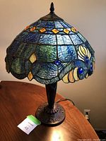 The lamp illuminated showing the stained glass shade in rich green and blue colors with amber accents and blue glass jewels.