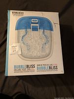 Front view of sealed box showing HoMedics Bubble Bliss Deluxe Foot Spa with blue top and control panel, showing the foot openings and bubble action illustration on the box.