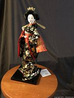 Full view of Japanese Geisha doll standing on black square base, showing full attire and posture.