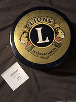 Closed view of vintage round Lions Club International tin with yellow and black design featuring two lions and a central letter 'L'.