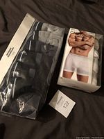 Photo showing both the Calvin Klein 5-pack boxer briefs package and Amazon Essentials 7-pack briefs package on a dark surface.