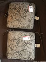 Two square cushions with black and grey tropical leaf design laid flat showing brand tags and ties for securing to chairs.