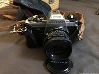 Front view of Pentax Super Program film camera with attached SMC Pentax-A 1:2 lens and Pentax lens cap, resting on a surface with leather carrying case strap visible.