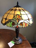 Front view of Tiffany style lamp showing stained glass shade with amber, green, orange accents and dark metal ornate base on wood table.