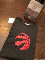 Folded dark gray men's long sleeved shirt with large red Toronto Raptors logo and accompanying clear display box with Coors Light and Raptors graphics.
