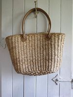 Front view of a natural straw market bag hanging on a wall showing woven texture and leather handle attachments.