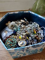 Top view of large floral patterned box filled with assorted costume jewelry pieces including pearl-like beads, turquoise, gold-tone jewelry, and mixed metal items