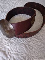 Wide brown leather belt coiled to show hammered metal buckle and part of perforated leather strap.