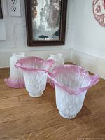 Four pink-rimmed white glass shades arranged on a wooden surface, showing flared ruffled tops and textured glass bodies.
