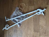 Set of two white painted metal swing curtain rods laid out on wooden floor with a plastic bag containing accessories.