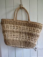 Front side of a two-toned woven straw market bag hanging on a white door, showing its braided handles and striped weaving pattern.