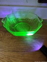 Photo showing octagonal uranium depression glass bowl glowing green under UV light on wooden surface, highlighting embossed floral patterns and two handles.