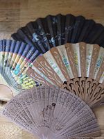 Four vintage hand fans arranged overlapping on wooden surface showing different styles and materials including black Victorian fabric with white flowers, colorful Asian paper, carved wood with painted flowers, and finely carved wood with painted silhouette.