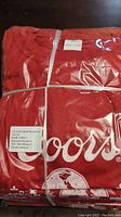 Photo showing folded red T-shirts with visible Coors Banquet branding through clear packaging.