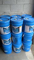 Stack of 18 blue 5-gallon pails with metal handles and white labels showing product name and brand, arranged three wide and six high.