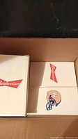 Cardboard box open showing three white boxes each with Budweiser branding and one with a football helmet logo.
