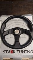 Full view of the Stark Tuning Type R steering wheel placed on the box showing the textured black grip and three-spoke design.
