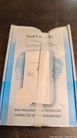 Front view of the packaged True Comfort floor warming system thermostat showing the product name and non-programmable feature, with a white strap securing the box.