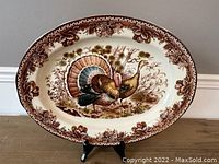 Full view of the large oval platter with turkey design and brown grapevine border against a beige background on a black stand.