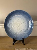 Full view of the large round ceramic plate with blue gradient and floral embossed design, displayed on a black stand against a beige wall.