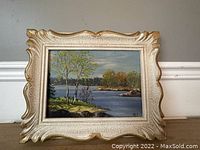 Framed landscape oil on board painting showing river scene with trees and rocks, signed, in ornate gold and cream frame.