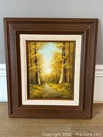 Full view of the framed oil on board painting showing a forest path with autumn colored trees, bright sky, and wooden brown frame with cream inner liner.