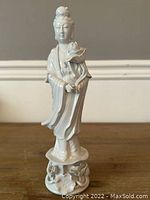 Full view of the Blanc De Chine Guanyin figurine showing the entire figure on a wooden surface against a gray wall, highlighting the white porcelain and detailed sculpting.