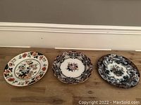 Three antique English Imari plates arranged side by side on a wooden surface, showing varying floral and abstract patterns with red, blue, black, and gold highlights.