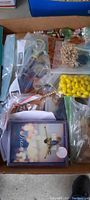 Box flat of mixed costume jewelry pieces in small plastic bags showing various colors and materials including yellow beads, metal chains, and boxed necklace.