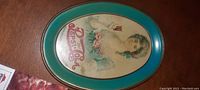 Oval tin tip tray with turquoise border depicting a woman holding a glass of Pepsi Cola and roses