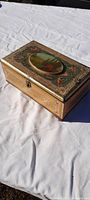 Top angled view of rectangular antique candy tin with hinged lid placed on white cloth outdoors, showing detailed tin litho landscape country scene on lid with floral border and gold accents.