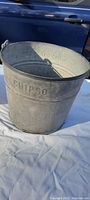 Front view of antique steel bucket showing embossed 'CHIPSO' lettering and embossed numbers inside the bucket, placed on white cloth with a blue vehicle in the background.