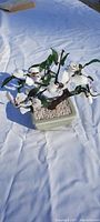 Artificial flower arrangement with white flowers and green leaves in light green ceramic pot on white surface.