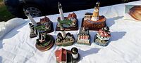 Overhead photo showing nine collectible lighthouse models with various architectural styles, colors, and attached building bases on white table cover.