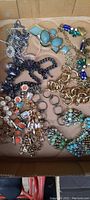Top view of assorted costume jewelry pieces in a box showing gold-tone chain bracelet, blue stone bracelet, multi-color earrings, and various other pieces.