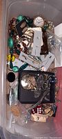 Photo showing plastic container filled with assorted pocket watches and costume jewelry including necklaces, earrings, and other small items.