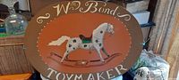 Full view of Wm Bond Toy Maker metal oval trade sign featuring a hand-painted white spotted rocking horse against a brownish-orange background with bordered edges.