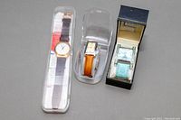 Overview of three ladies watches in their boxes or containers: a round gold-tone Swatch, rectangular AleniaAeronautica, and square rhinestone accented Collezio watch.