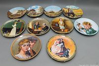 Ten vintage porcelain collectible wall plates arranged on grey surface showing artwork featuring scenic villages, horse-drawn carriages, and human figures.