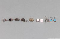Five pairs of 925 silver earrings arranged side by side showing different styles including heart-shaped, gemstone dangles, square studs, and opal-like stones.