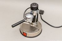 Side profile view of the espresso machine showing stainless base, chrome espresso head with black handle, power cord, and red switch.