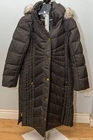 Front view of a black ladies long winter coat by Anne Klein, size XL, hanging on a stand. Quilted design with gold-tone snap buttons, hood with faux fur trim visible.