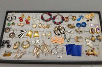 Top-down photograph of a black tray containing over 25 pairs of assorted vintage costume earrings featuring a wide variety of styles, colors, and shapes including hoops, studs, dangles, floral, and geometric designs.