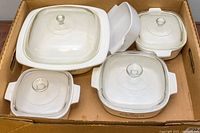 Five vintage CorningWare casserole dishes with lids visible, packed in a cardboard box showing some wear on surfaces.
